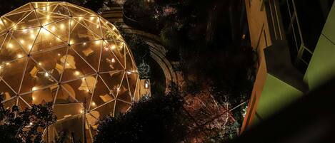 Estructura en forma de cúpula geométrica con asientos en su interior, rodeada de árboles y un sendero de ladrillos.