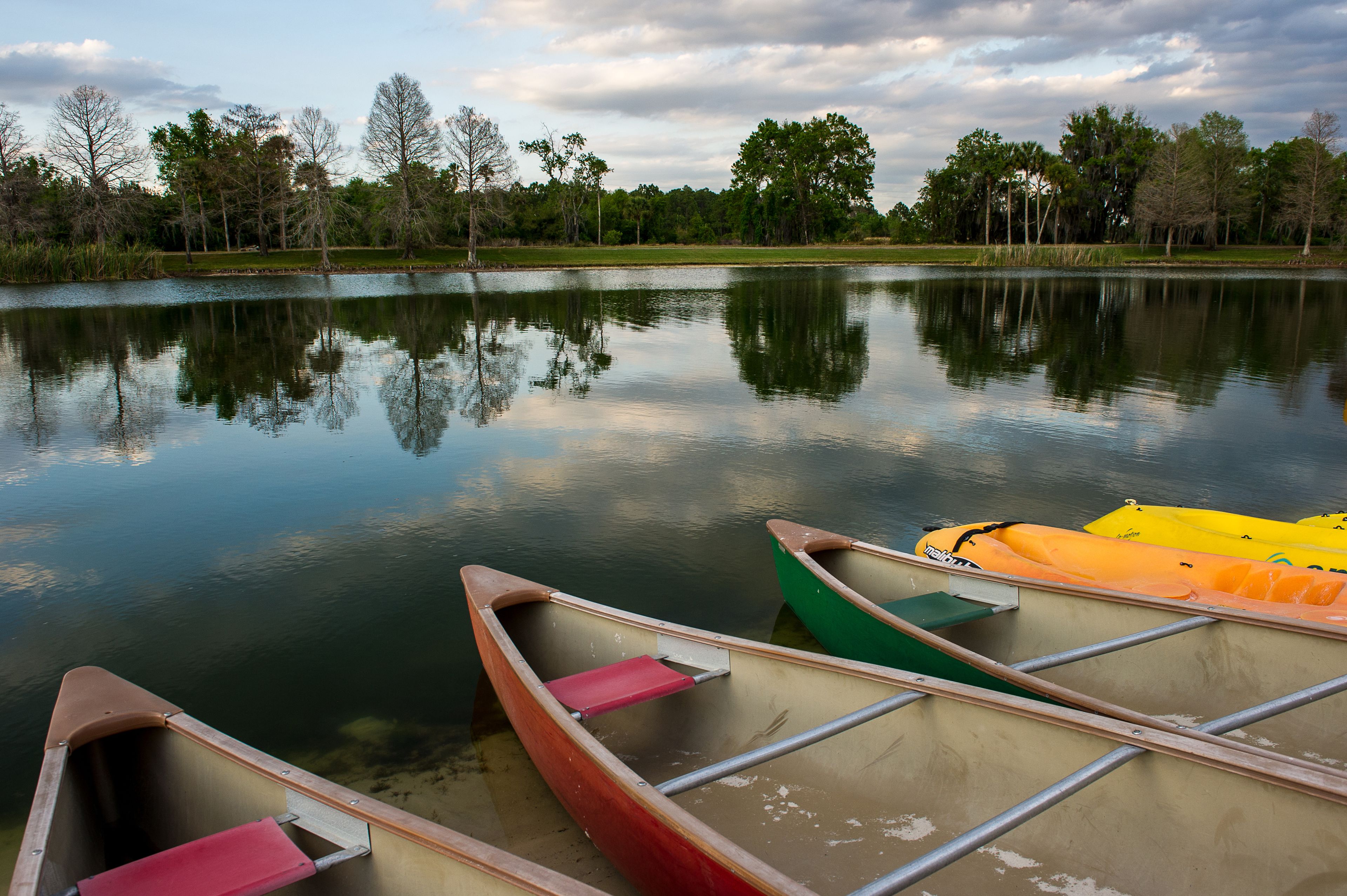 A calm lake with reflections of trees and a cloudy sky. There are several canoes and kayaks docked along the shore.