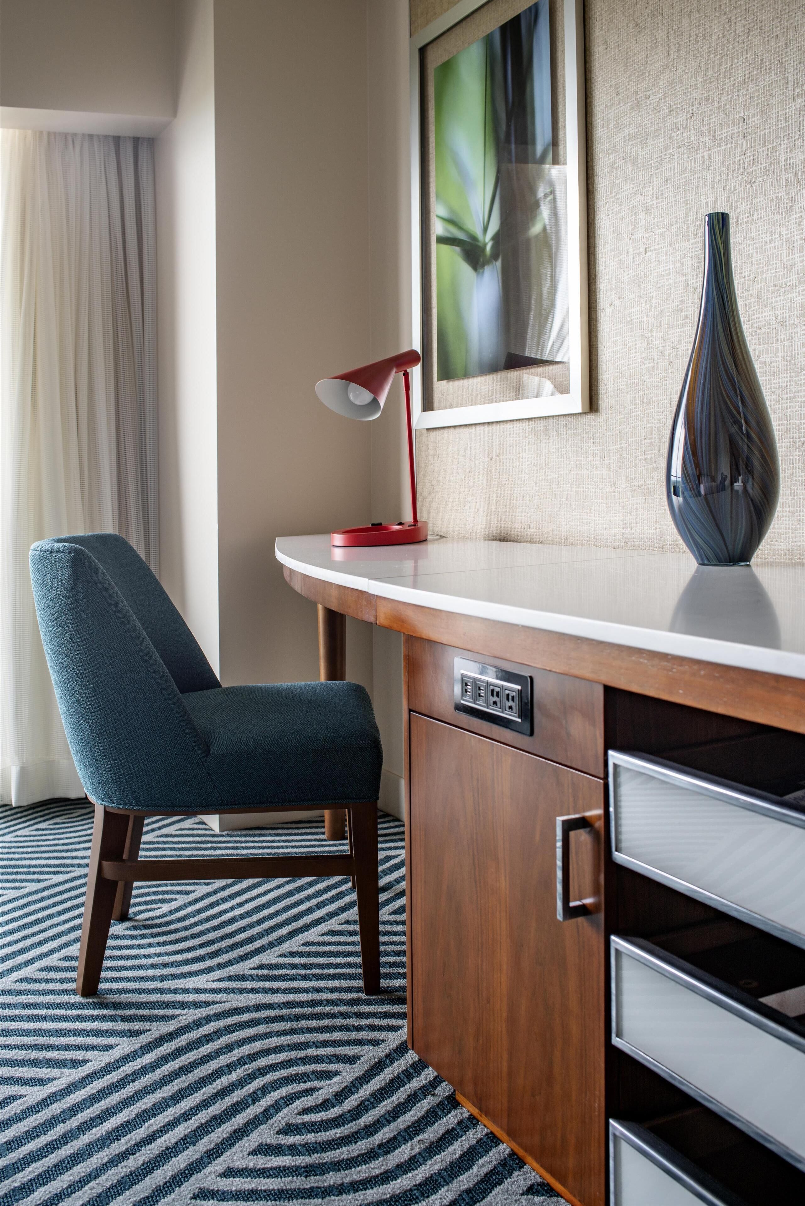A hotel room with a wooden desk, a blue chair, a red desk lamp, and a framed picture on the wall.