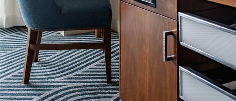 A hotel room with a wooden desk, a blue chair, a red desk lamp, and a framed picture on the wall.