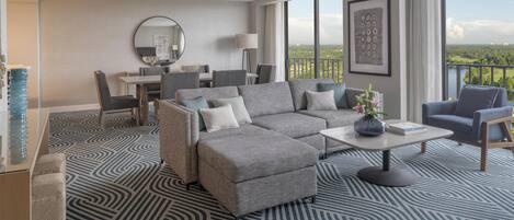 Una habitación de hotel moderna con un sofá seccional gris, una mesa de centro redonda y un área de comedor con mesa y sillas.