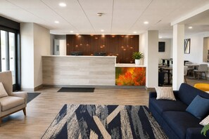 A reception area with a wooden reception desk, a potted plant, and a colorful autumn leaf wall. There are seating areas with sofas and a dining table in the background.