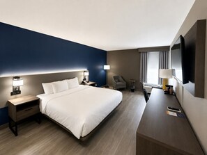 Ein Hotelzimmer mit einem großen Bett, Nachttischen, einem Schreibtisch, einem Sessel, einem Fernseher und einem fenster mit Vorhängen.