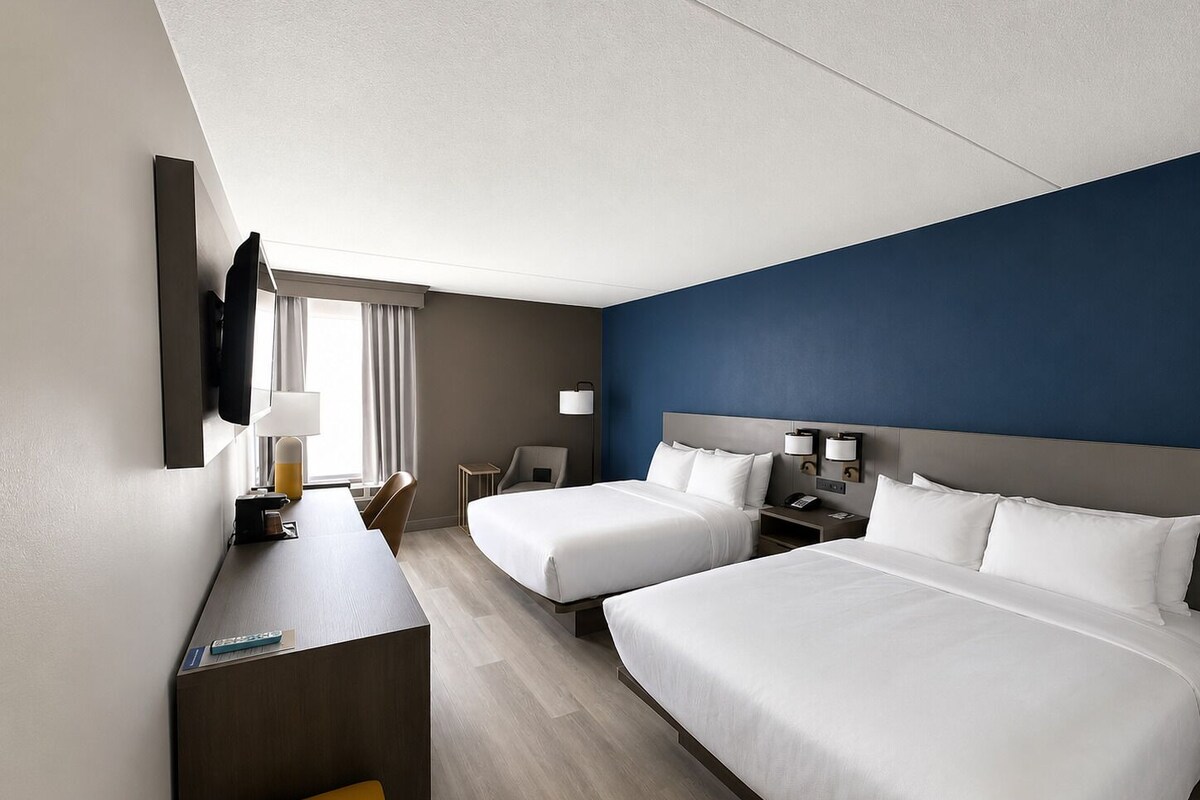 Ein Hotelzimmer mit zwei Betten, einem Schreibtisch, einem Stuhl, einem Fernseher und einem Fenster mit Vorhängen.