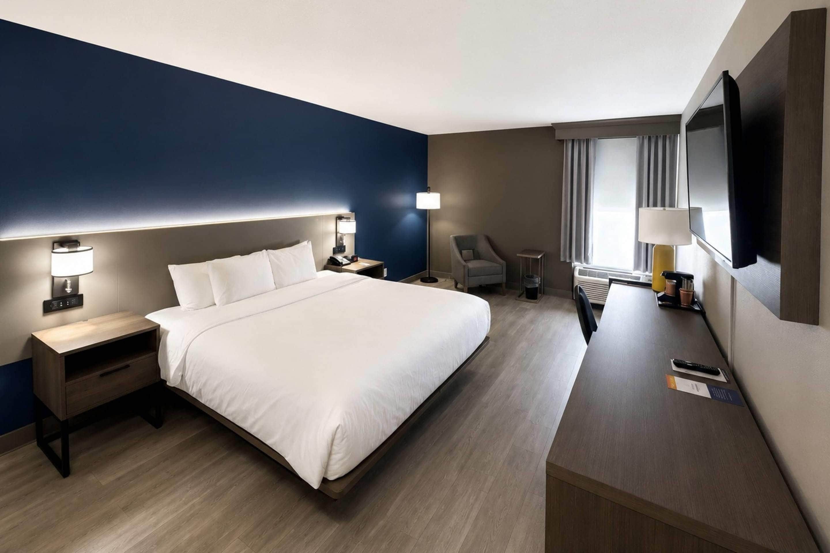 A hotel room with a large bed, bedside tables, a desk, a chair, a TV, and a window with curtains.