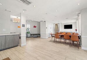 A modern hotel lobby with a reception desk, a dining area with orange chairs, and a flat-screen TV mounted on the wall.