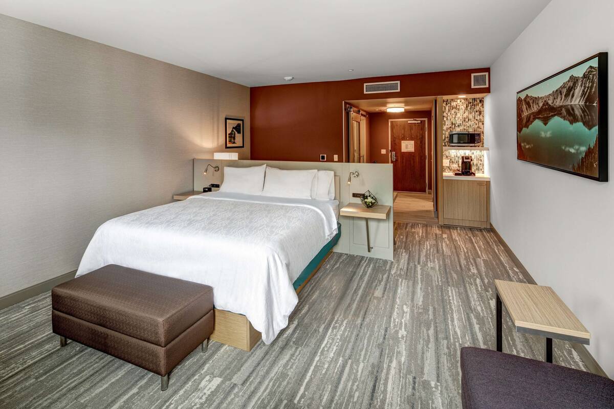 A hotel room with a large bed, a small table, a bench, and a view of a mountain through a window.