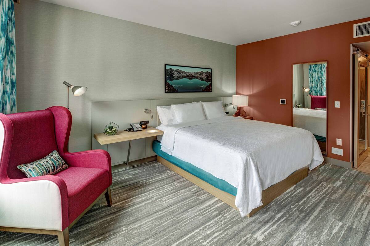 A hotel room with a bed, a desk, a chair, a nightstand, and a painting on the wall.