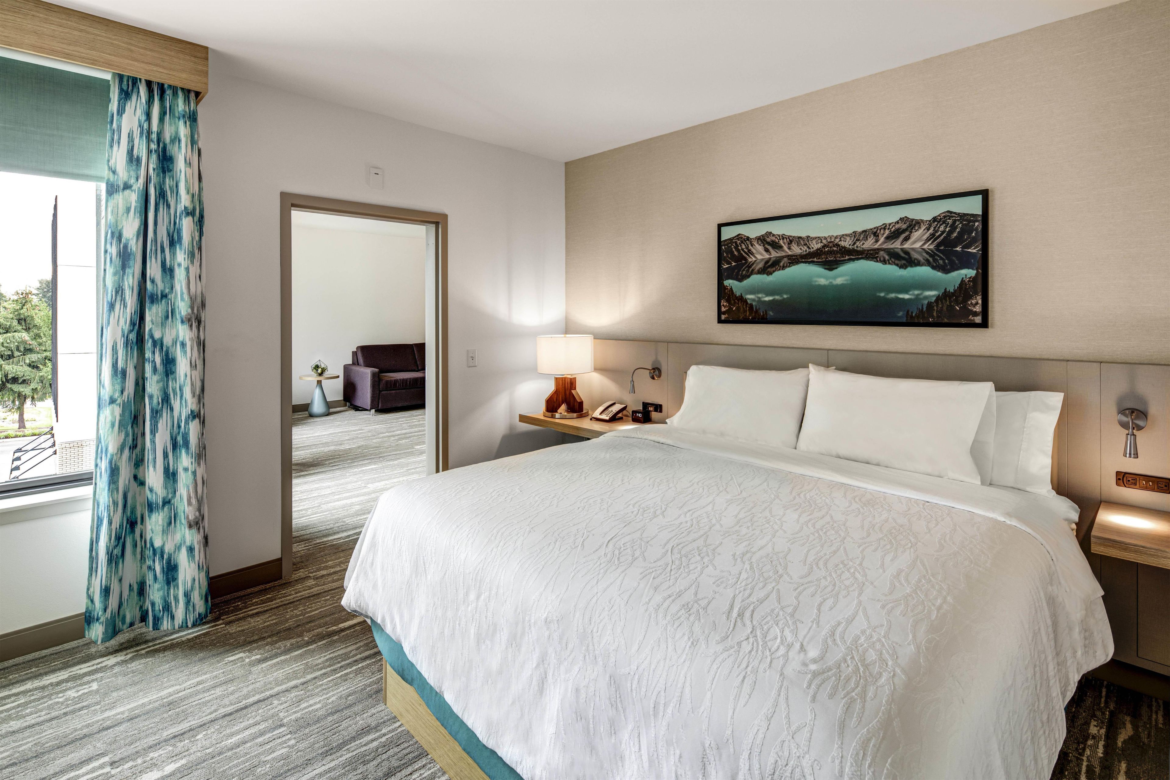 A hotel room with a large bed, a nightstand with a lamp, a painting of a mountain landscape, and a view of a balcony with a blue curtain.