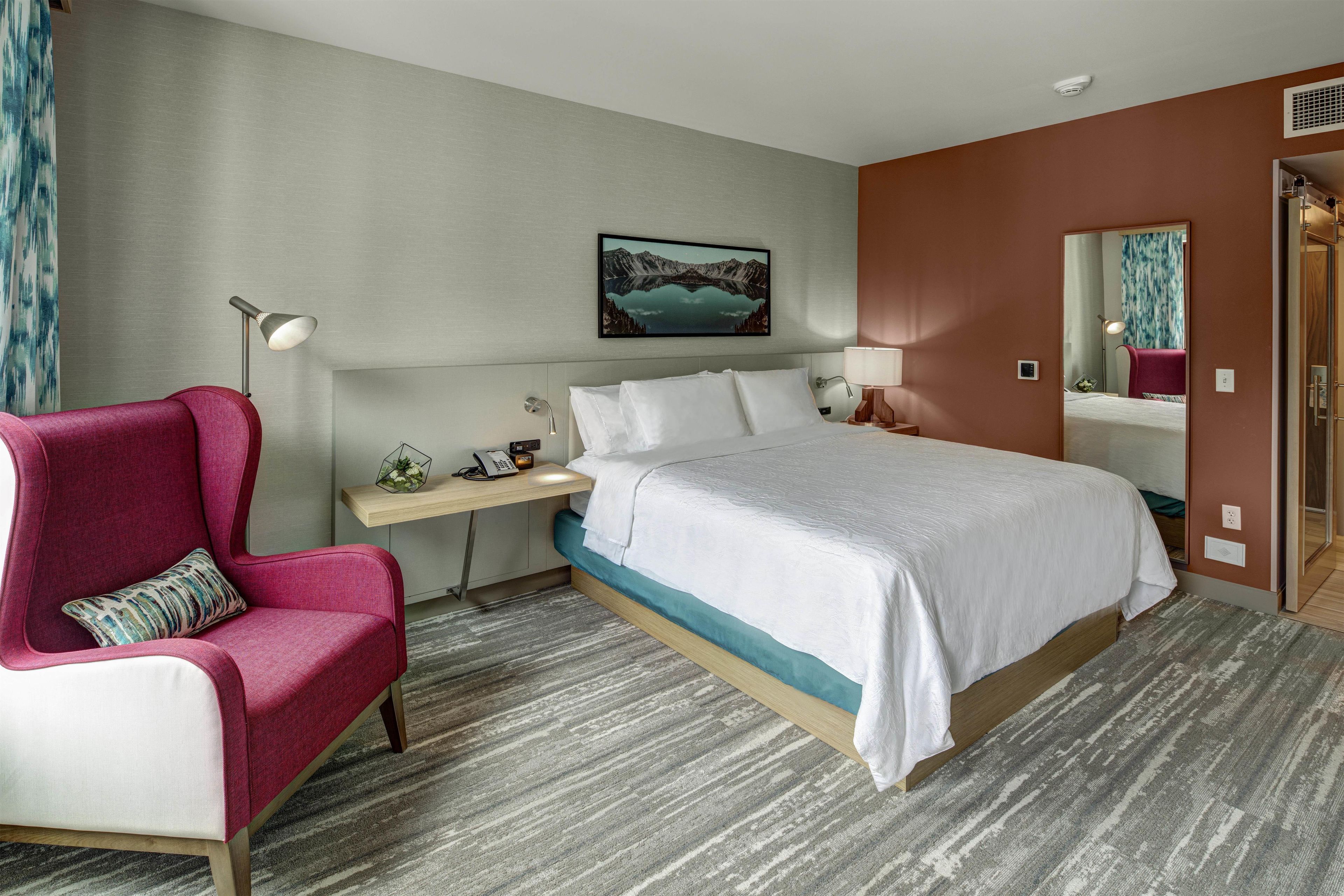 A hotel room with a bed, a desk, a chair, a nightstand, a lamp, a picture on the wall, and a mirror.