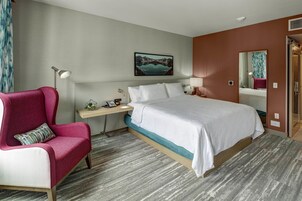 Habitación de hotel con cama, escritorio, silla, mesita de noche, lámpara, un cuadro en la pared y espejo.