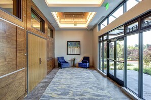 A hallway with wooden cabinets, two blue chairs, a painting on the wall, and large windows with a view of the outdoors. - Hilton Garden Inn Wilsonville Portland (Wilsonville)