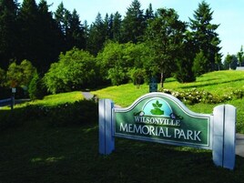 Señal para Wilsonville Memorial Park con una combinación de colores verde y blanco, rodeada de exuberante vegetación y árboles.