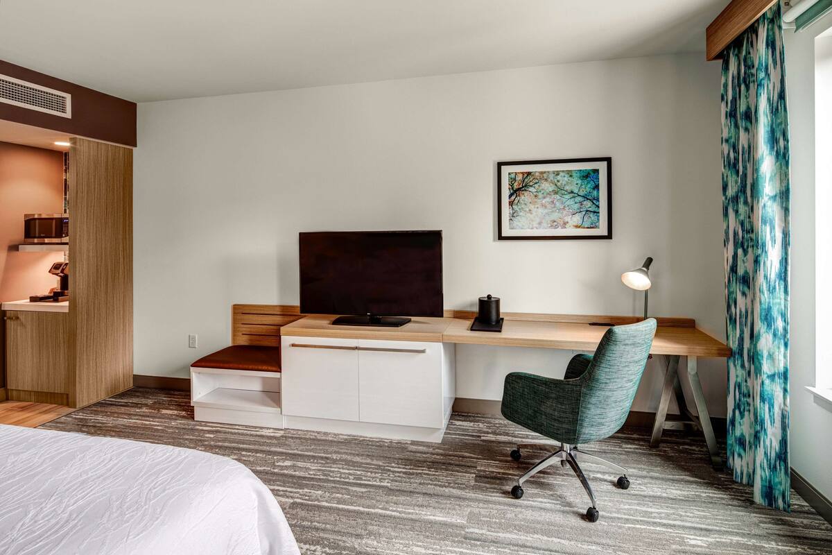A hotel room with a bed, a desk with a computer, a TV, and a framed picture on the wall.
