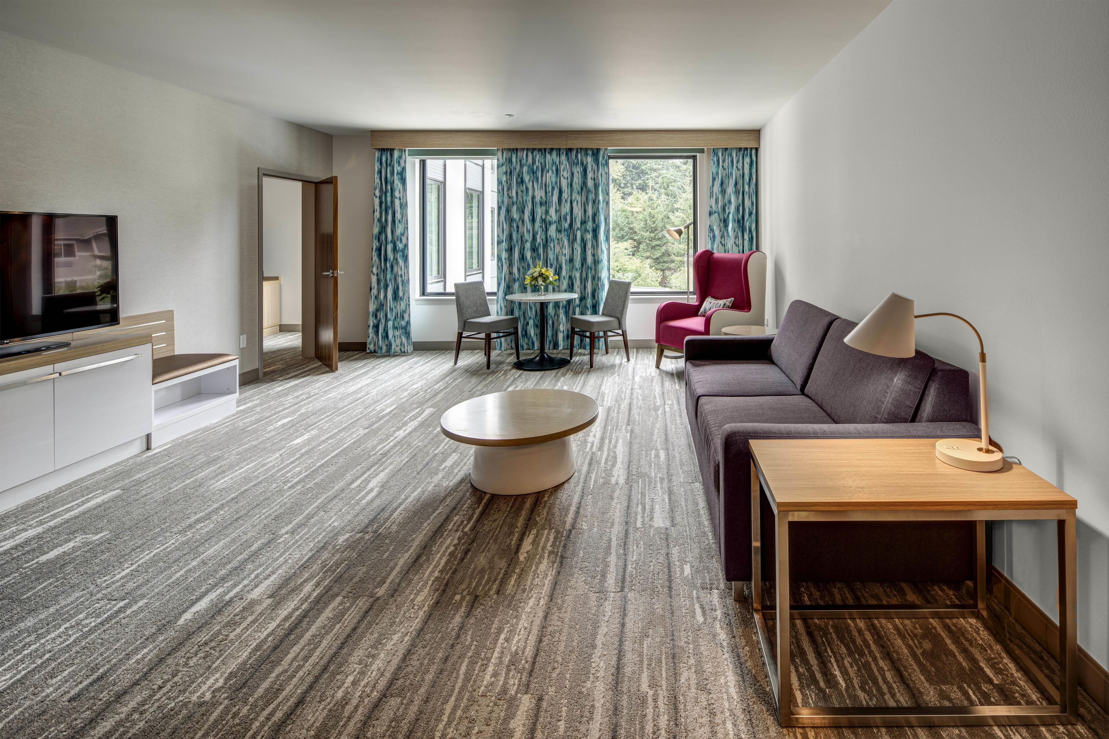 A modern hotel room with a flat-screen TV, a round coffee table, a sofa, a small side table, and a lamp.
