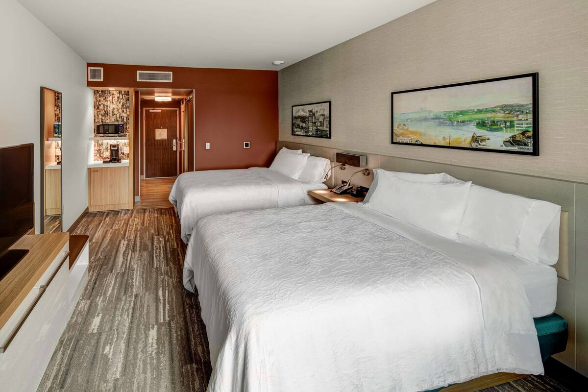 A hotel room with two beds, a kitchenette, and a painting of a landscape on the wall.