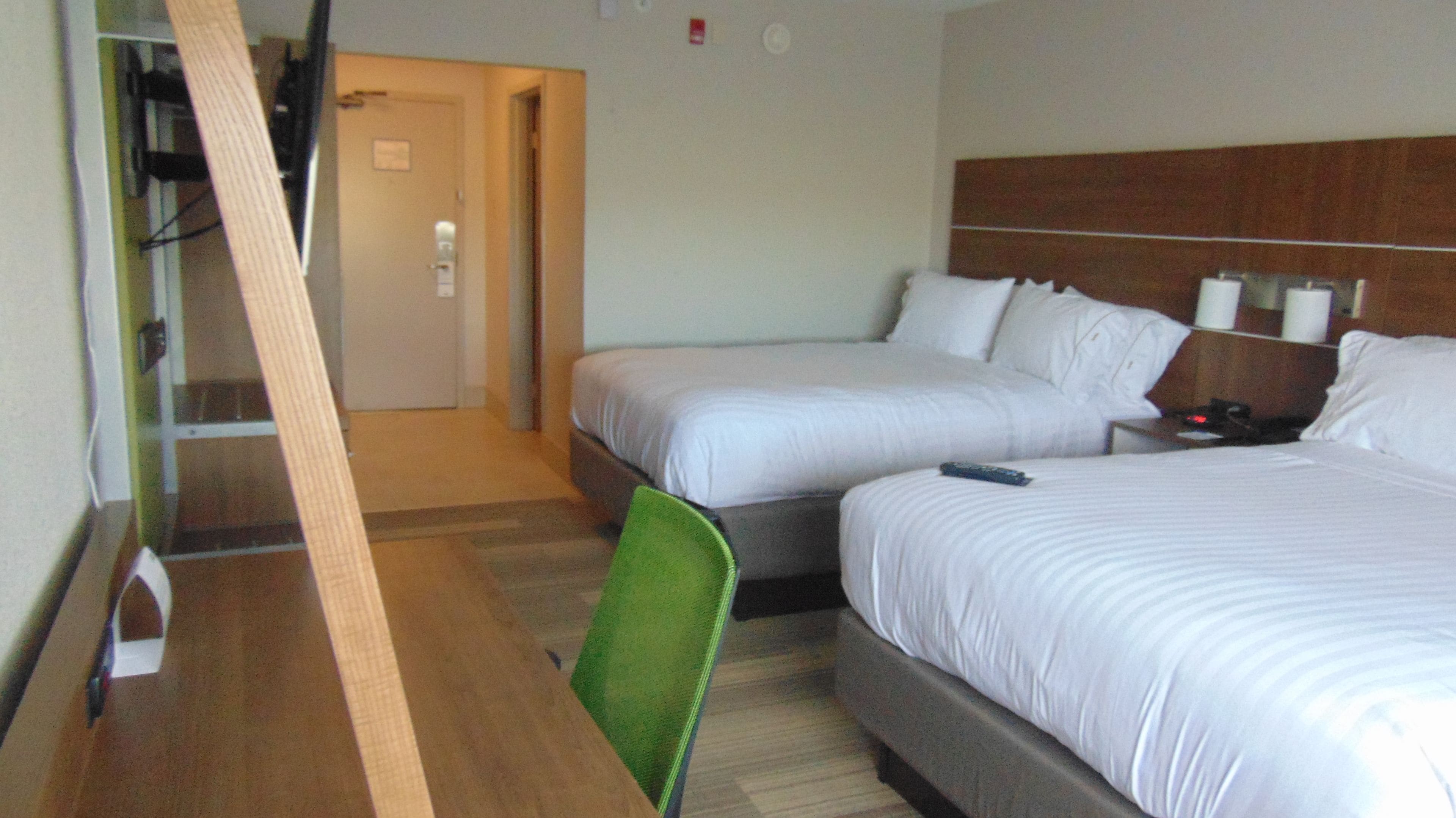 A hotel room with two beds, a wooden desk, a green chair, and a nightstand with a remote control.