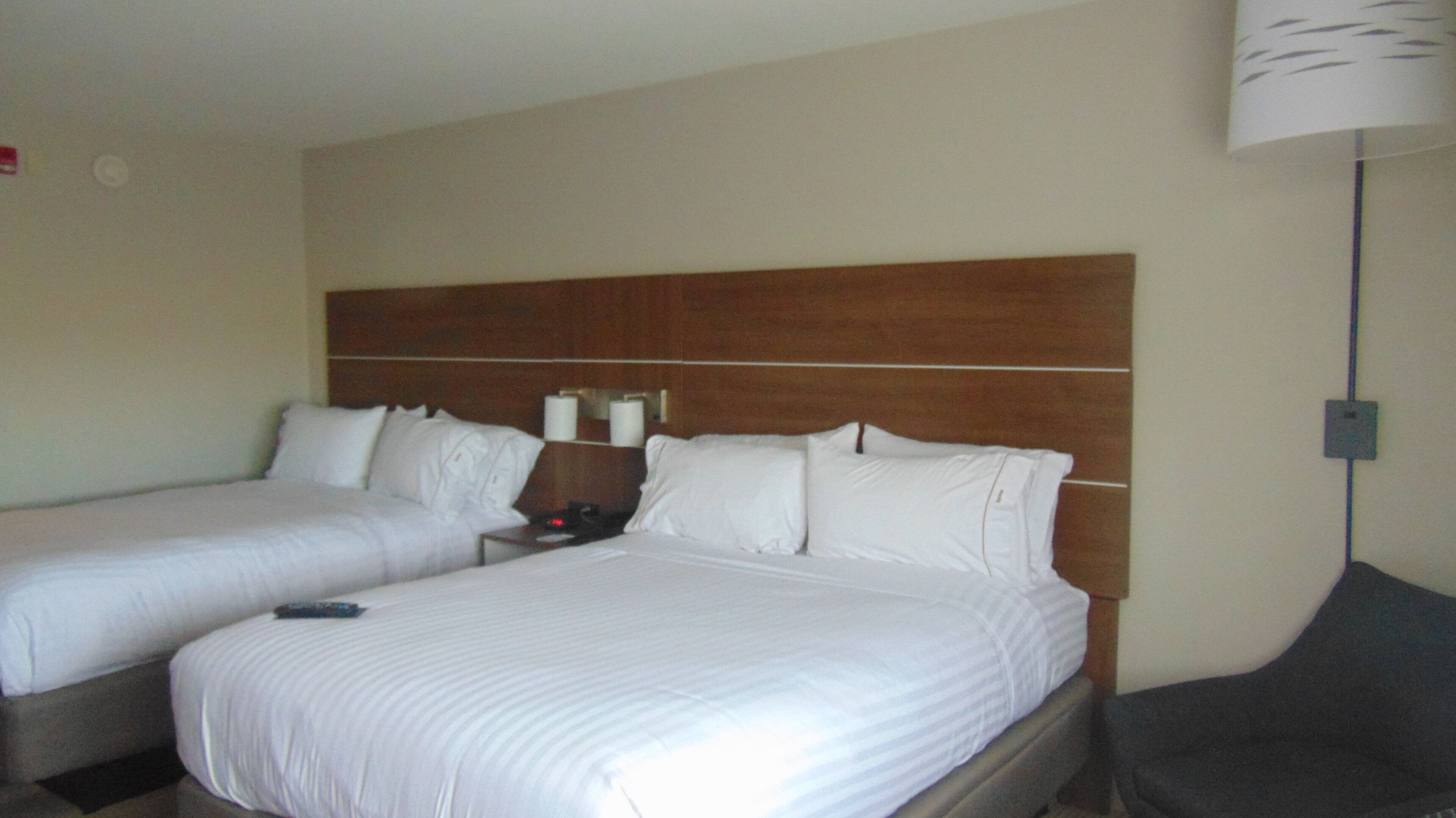 Habitación de hotel con dos camas individuales, cabecera de madera, mesita de noche con lámpara y un enchufe en la pared.