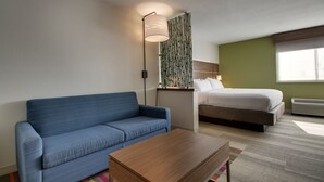 Une chambre d’hôtel avec un canapé bleu, une table basse en bois, un lit avec une literie blanche, une lampe fixée au mur et une fenêtre laissant entrer la lumière naturelle.