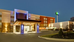 Un hôtel moderne doté d’une entrée bien éclairée, d’une enseigne bien visible indiquant qu’il s’agit d’un Holiday Inn Express, et d’un espace paysager agrémenté d’un banc et d’un petit arbre.