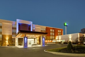 A modern hotel with a well-lit entrance, a visible sign indicating it's a Holiday Inn Express, and a landscaped area with a bench and a small tree. - Holiday Inn Express & Suites Wapakoneta by IHG (Wapakoneta)