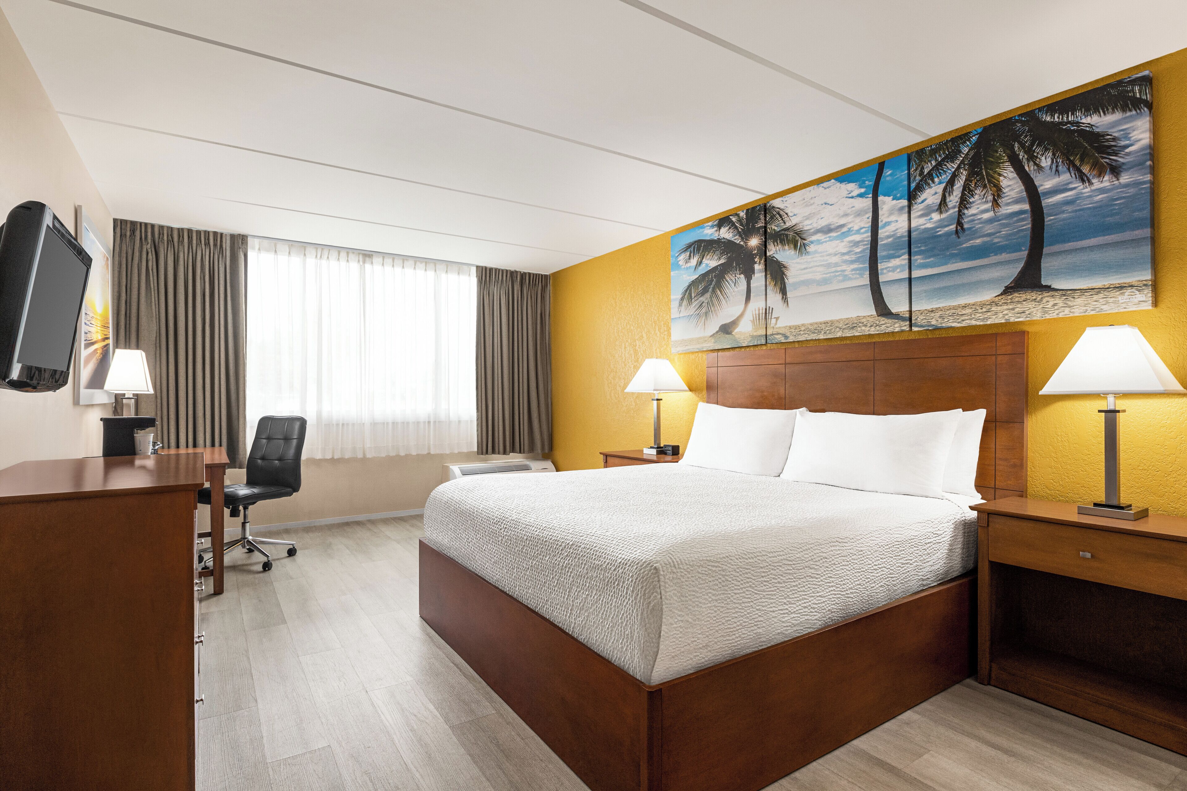 A hotel room with a bed, desk, chair, and a painting of palm trees on the wall.
