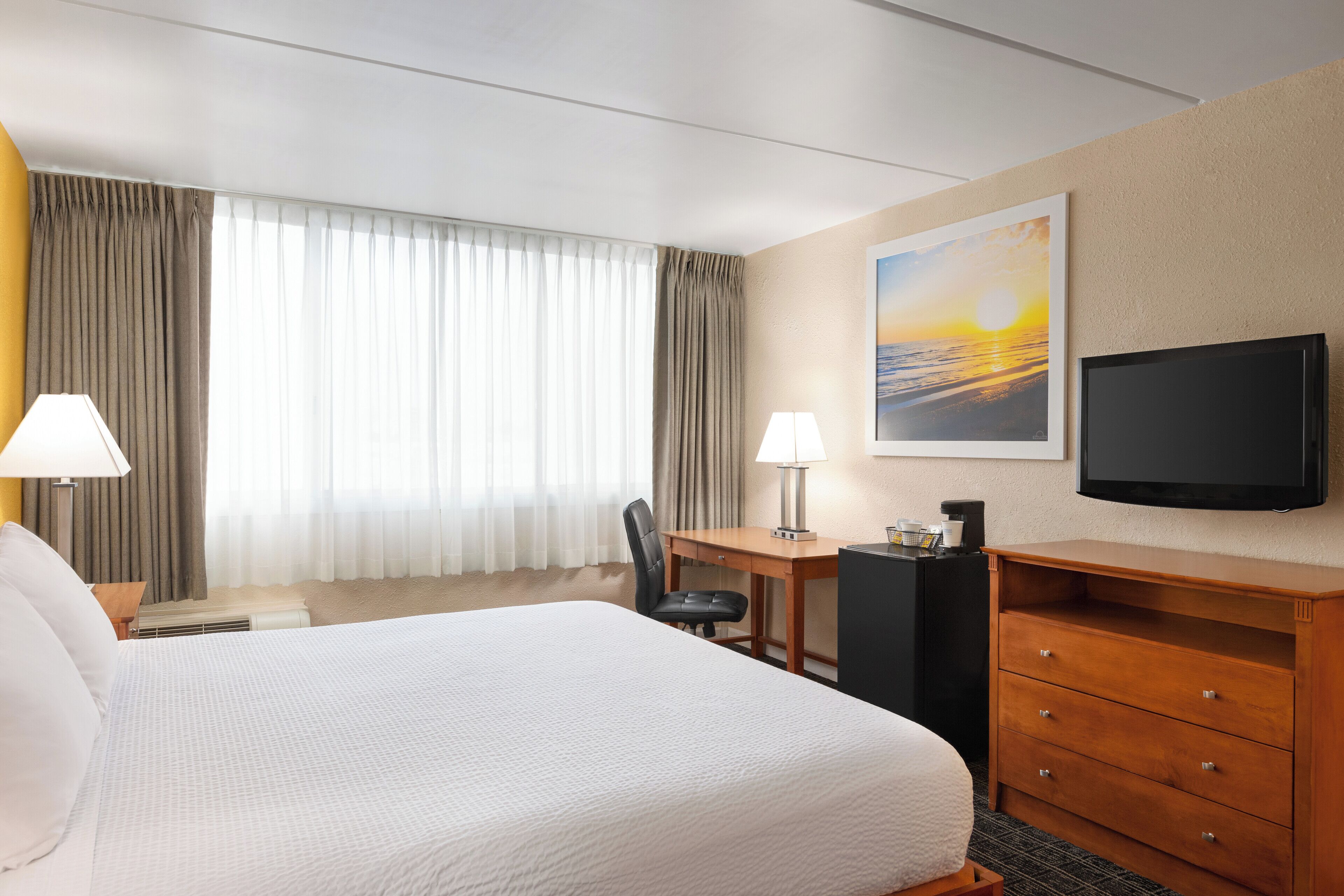 A hotel room with a large bed, a desk with a chair, a television, and a painting of a sunset.