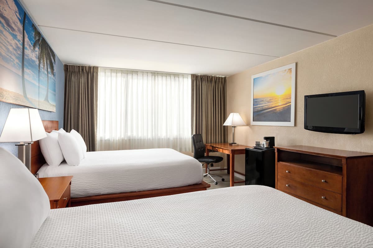 A hotel room with two beds, a desk, a chair, a television, and a painting of a sunset.