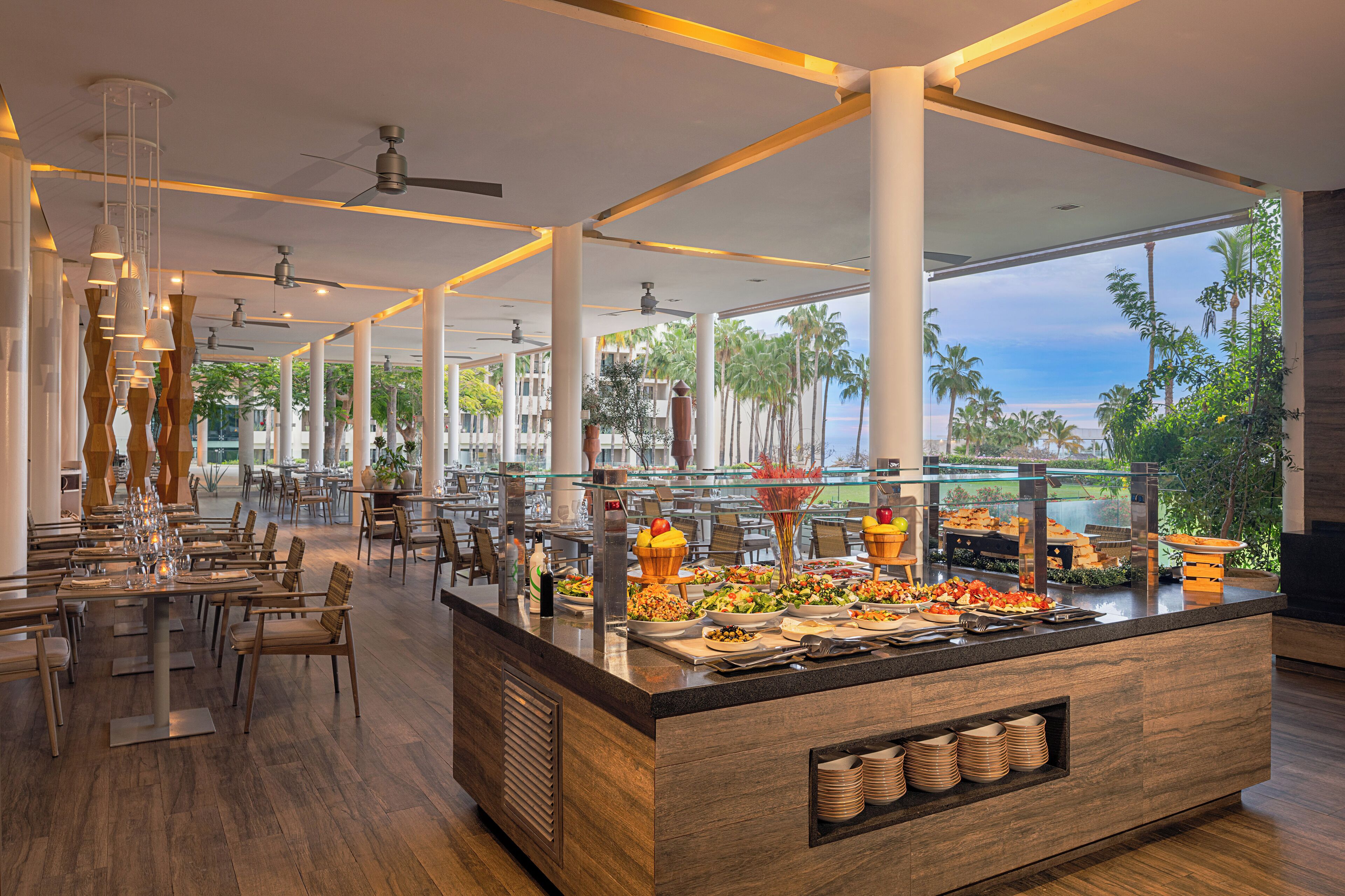 Uma sala de jantar moderna com buffet, mesas e cadeiras de madeira, amplas janelas com vista para palmeiras e o oceano.