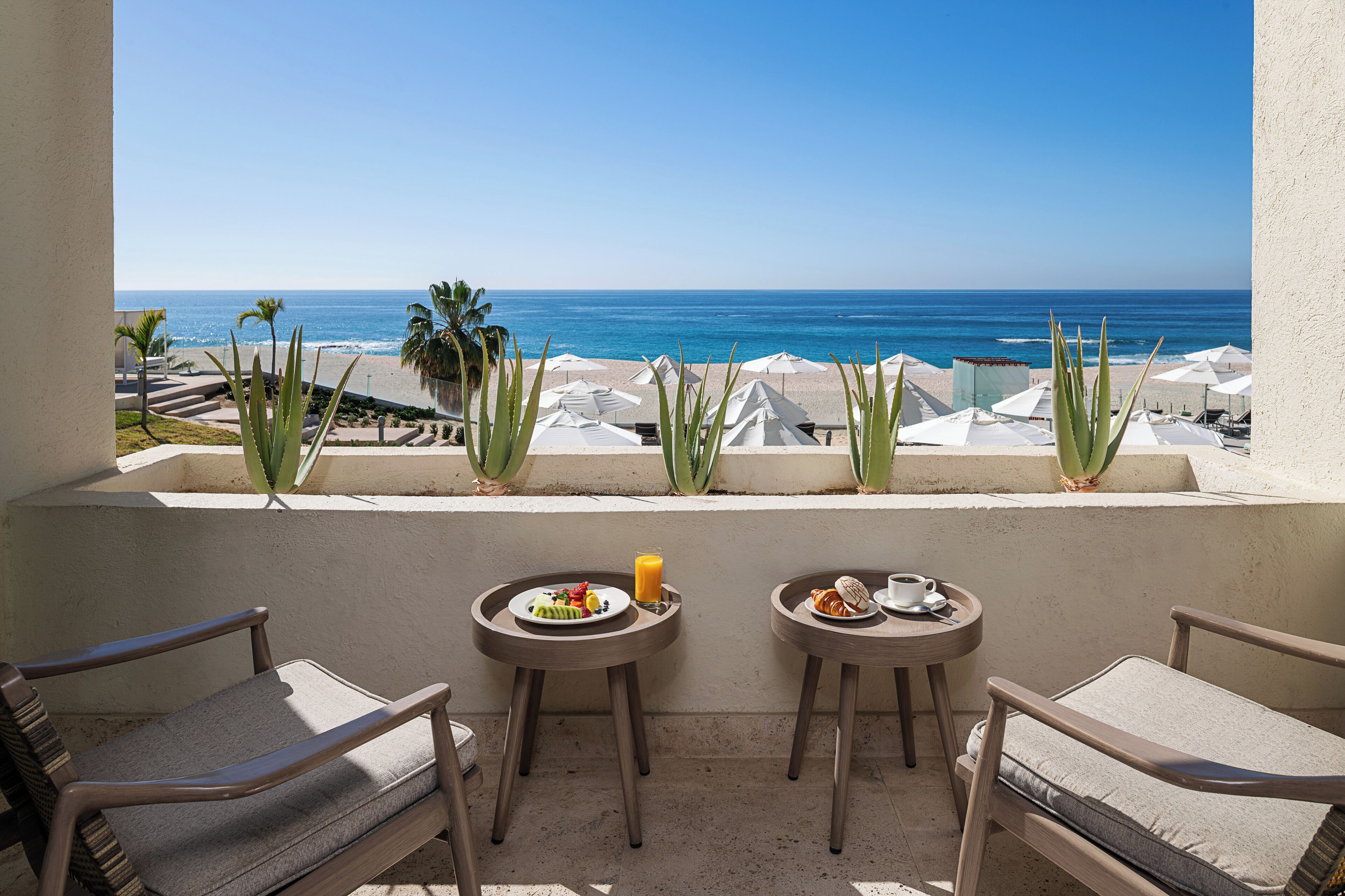 A balcony with two chairs and a table, overlooking a beach with umbrellas and palm trees.