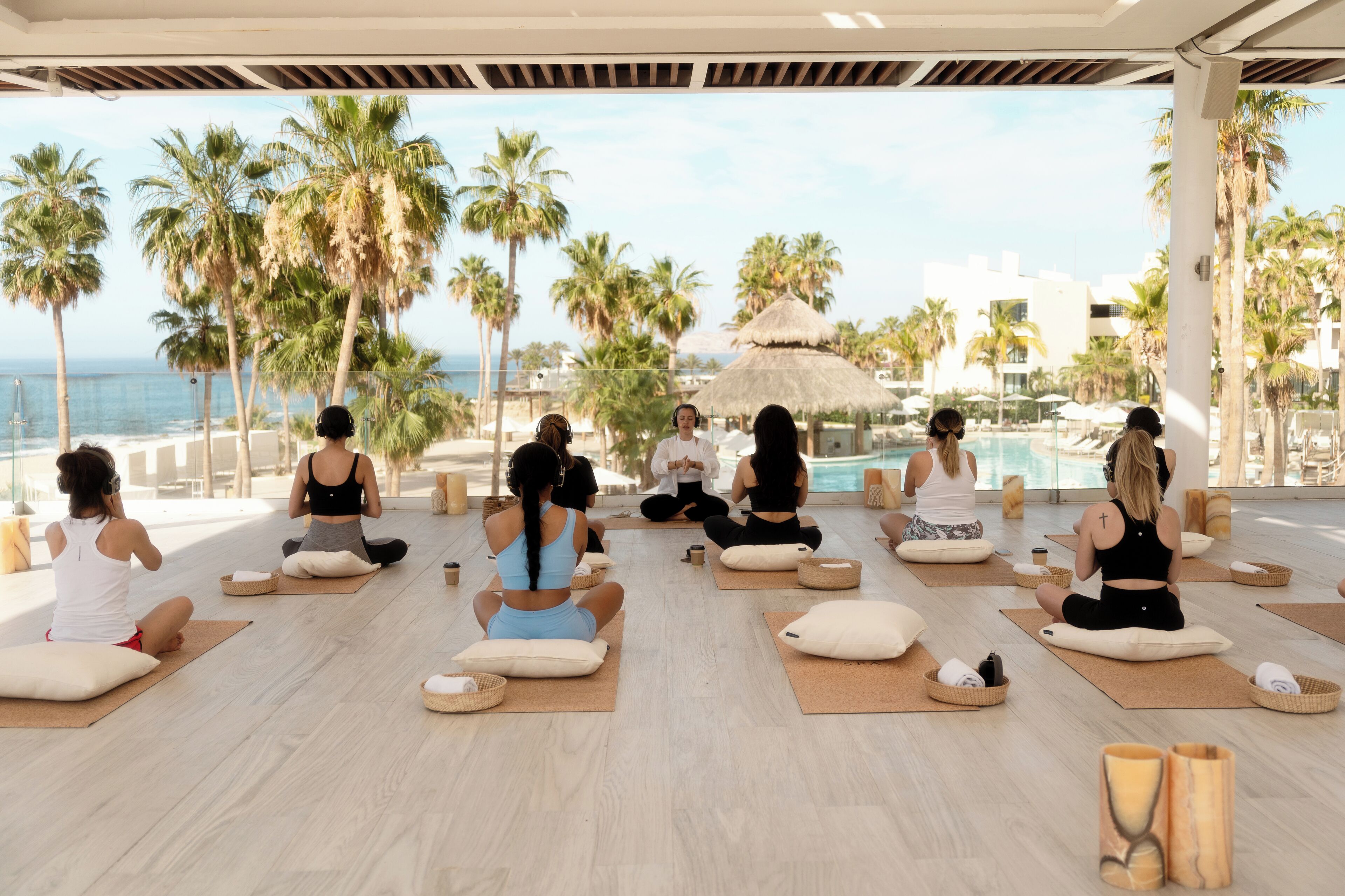 A group of people sitting on yoga mats in a circular arrangement on a wooden floor, with a pool and palm trees in the background.