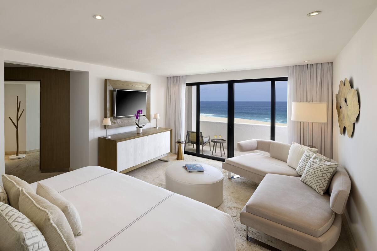 Ein Hotelzimmer mit einem großen Bett, einer Sitzecke, einem Fernseher, einem Balkon mit Blick auf den Strand und einem kleinen Tisch mit zwei Stühlen.