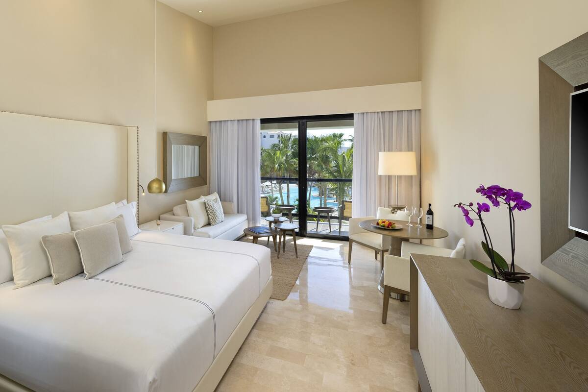 Ein Hotelzimmer mit einem großen Bett, einer kleinen Couch, einem Esstisch mit Stühlen, einem Fernseher und Blick ins Freie.