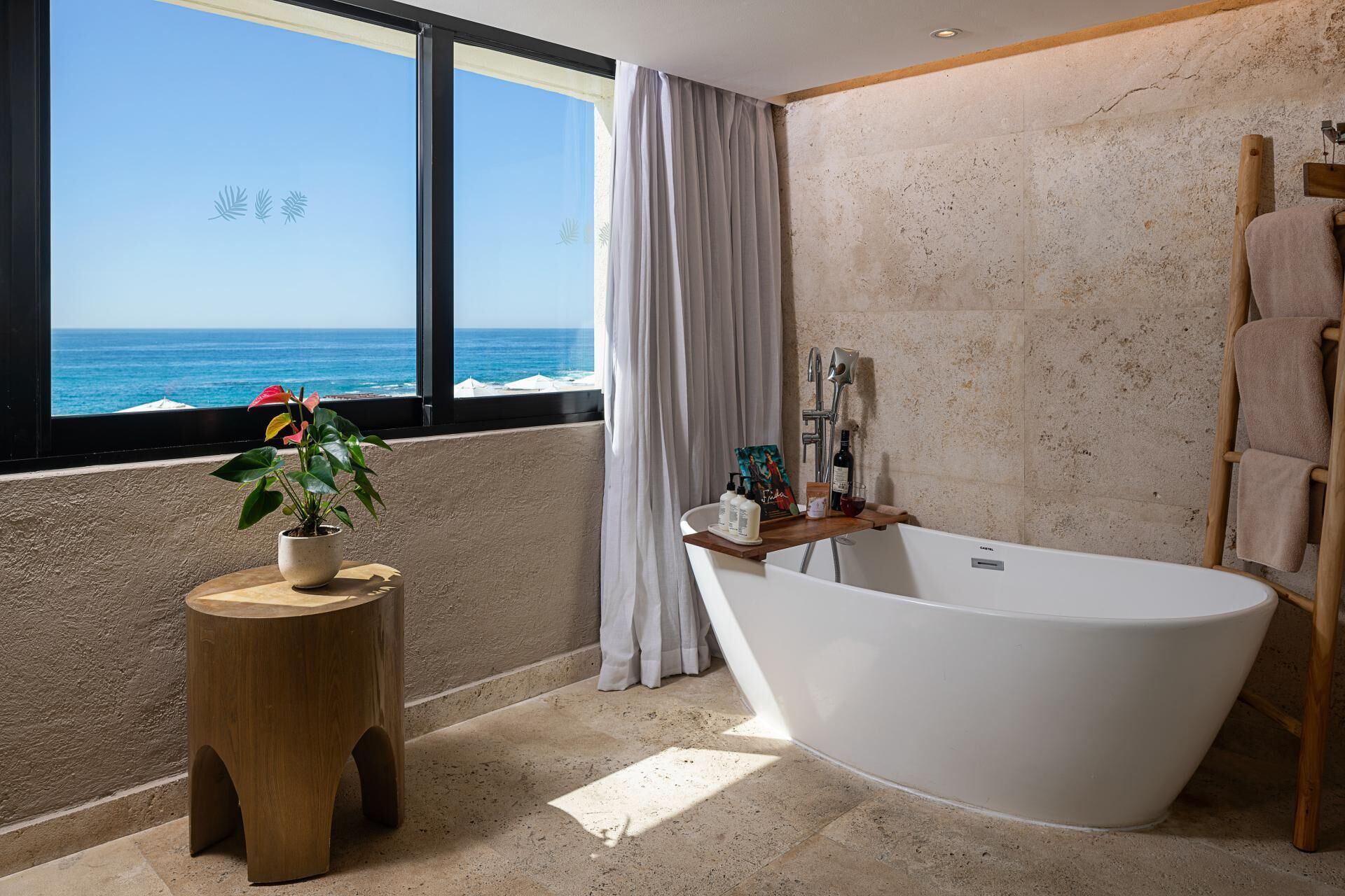 Un baño moderno con una bañera independiente, un taburete de madera con una planta y vistas al océano a través de amplias ventanas.