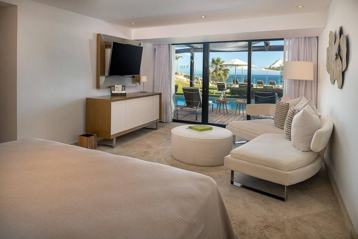 Ein Hotelzimmer mit Bett, einer Sitzecke mit Blick auf Pool und Meer sowie einem an der Wand montierten Fernseher.