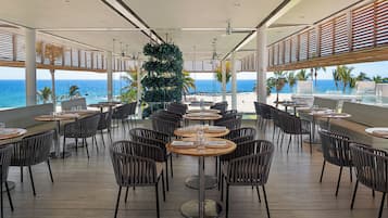 A restaurant with round tables and chairs, a view of the ocean, and a large tree in the center.