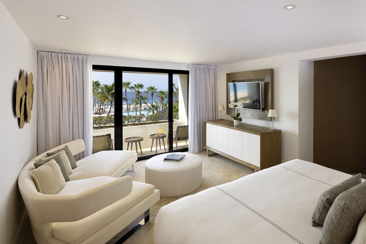 A hotel room with a bed, a sofa, a desk, a TV, and a balcony with a view of the beach.
