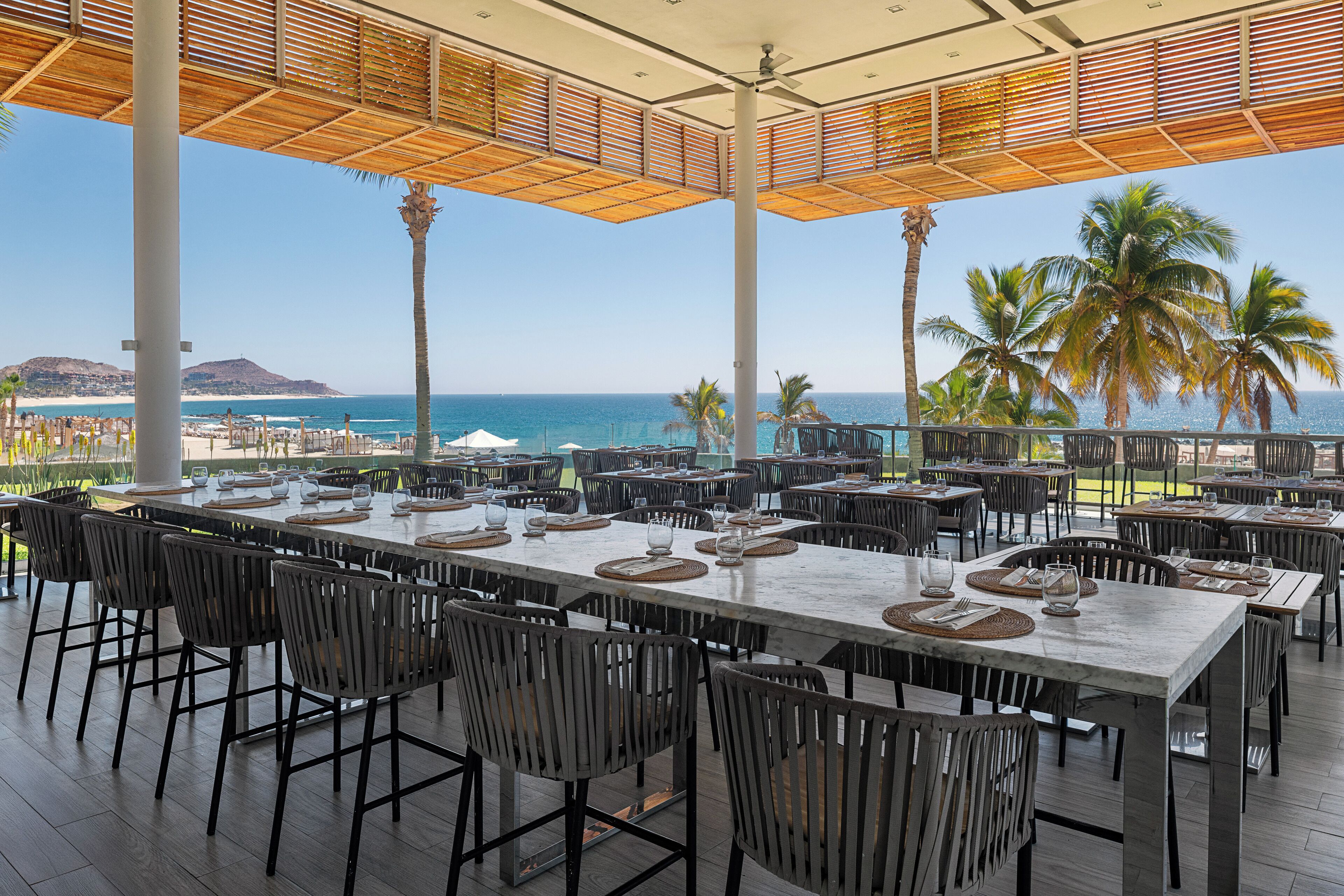 An outdoor dining area with multiple tables and chairs, overlooking a beach and ocean.