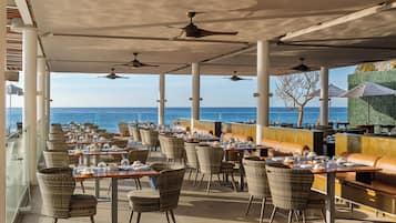 An outdoor dining area with tables and chairs, ceiling fans, and a view of the ocean.