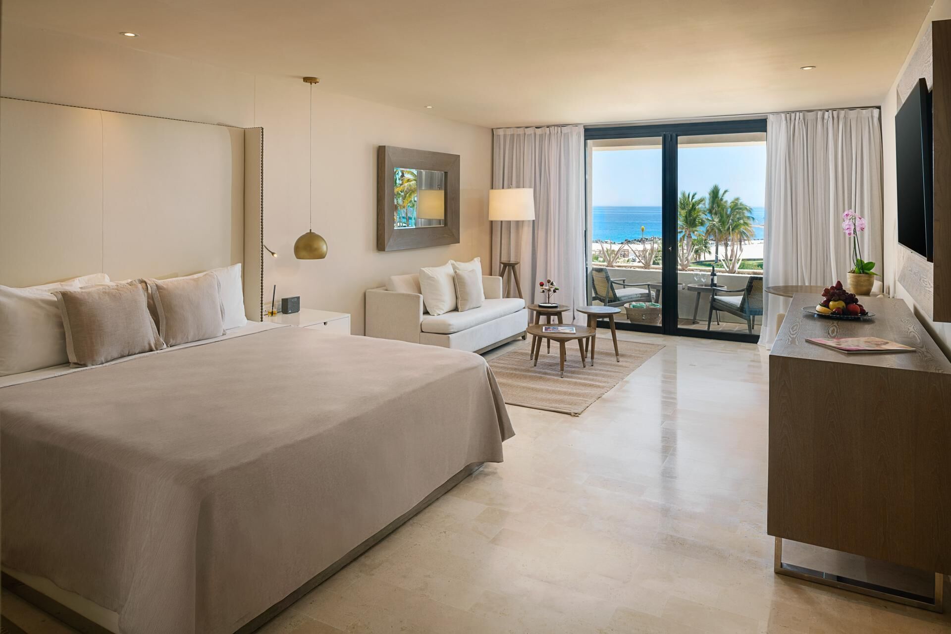 A hotel room with a large bed, a sofa, a dining area with a table and chairs, and a balcony with a view of the ocean.
