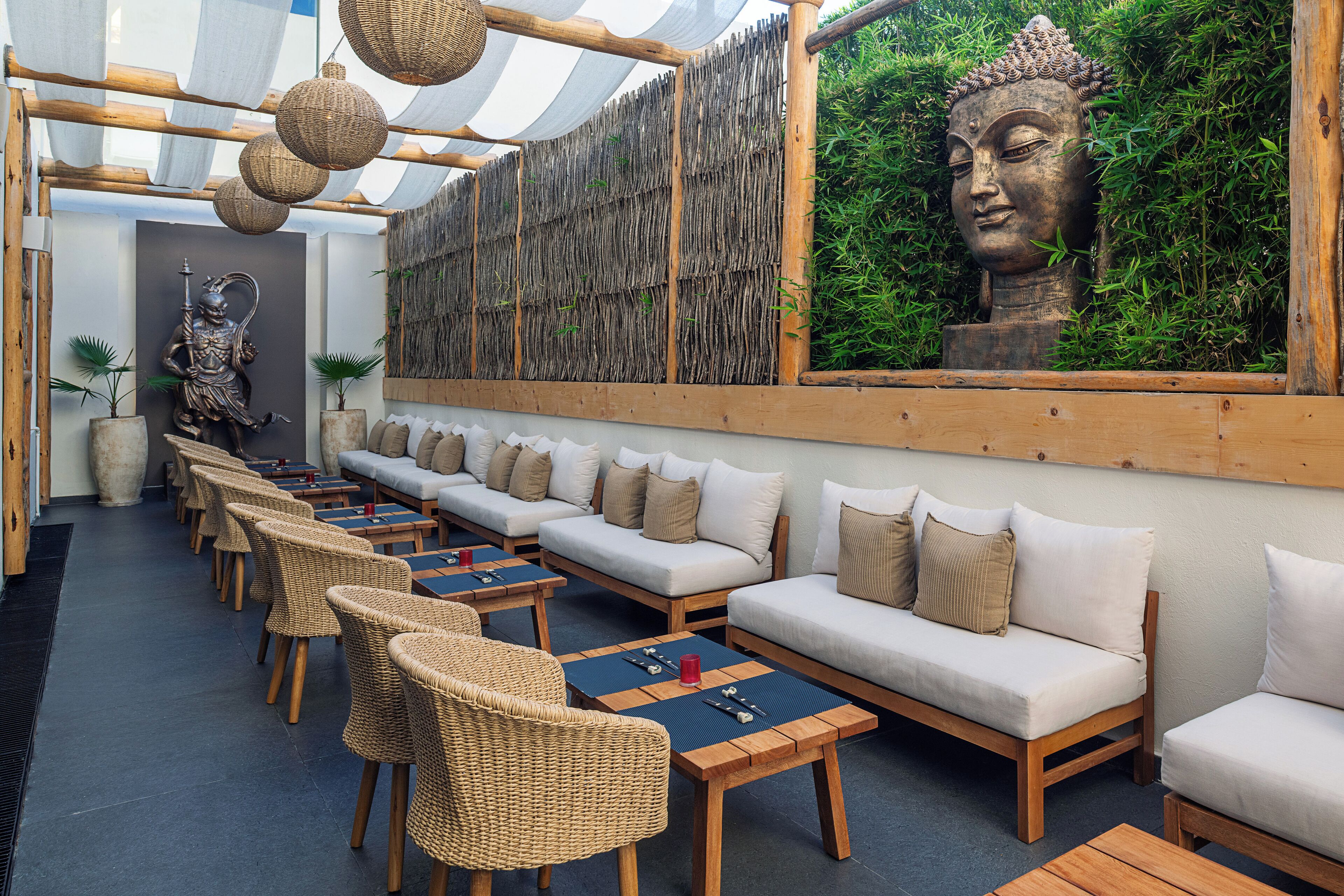 A restaurant with a long row of wicker chairs and sofas, a Buddha statue, and a bamboo wall.