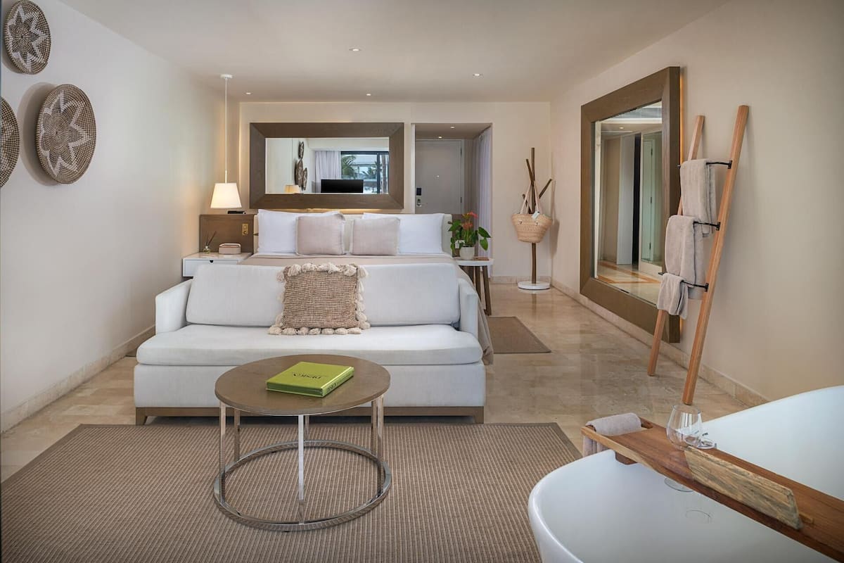 Ein modernes Hotelzimmer mit einer Couch, einem kleinen runden Tisch, einem Bett mit Blick ins Freie und einem großen Spiegel.