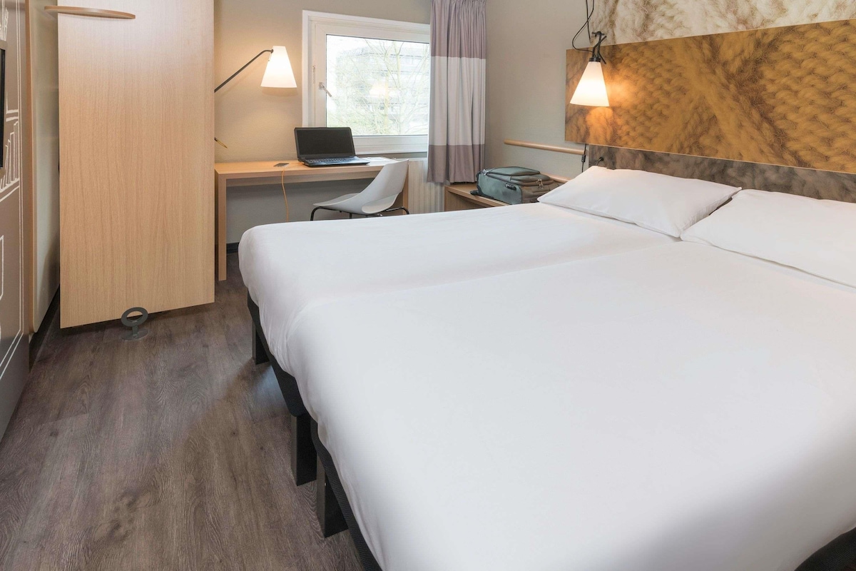 A hotel room with a bed, a desk with a laptop, a chair, and a nightstand with a lamp.