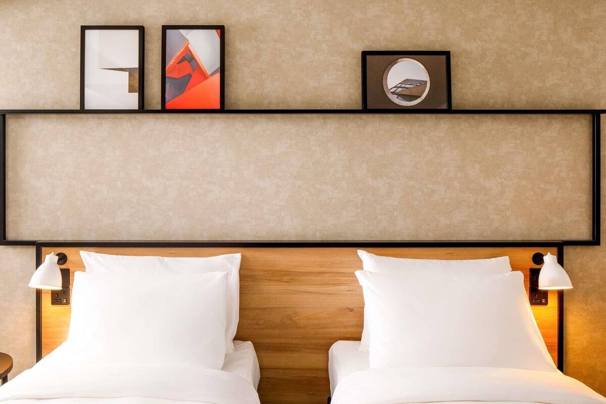 A hotel room with a wooden headboard, two beds, white bedding, a wall-mounted shelf with framed artwork, and bedside lamps.