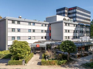 A modern hotel with a central entrance, outdoor seating area, and a parking lot. - ibis Tilburg (Tilburg)