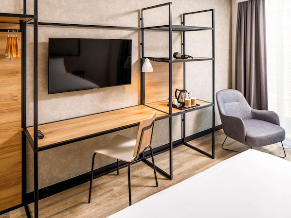 A modern hotel room with a wooden desk, a grey armchair, a TV mounted on the wall, and a shelf with a lamp and a coffee maker. - ibis Tilburg (Tilburg)