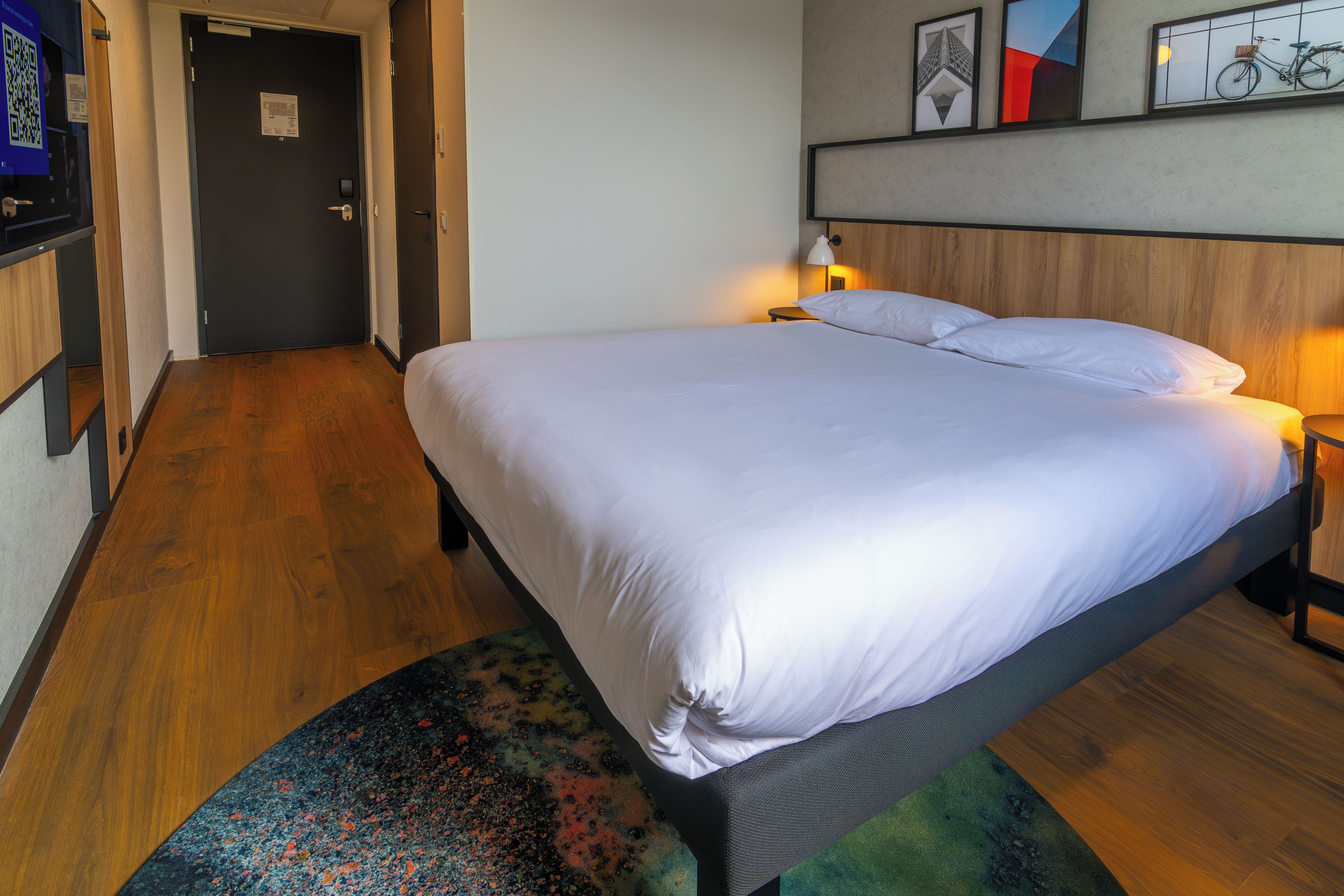 Una habitación de hotel con cama, mesita de noche, lámpara, cabecera de madera, una puerta y un cuadro en la pared.