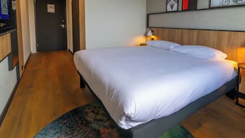 Una habitación de hotel con cama, mesita de noche, lámpara, cabecera de madera, una puerta y un cuadro en la pared.