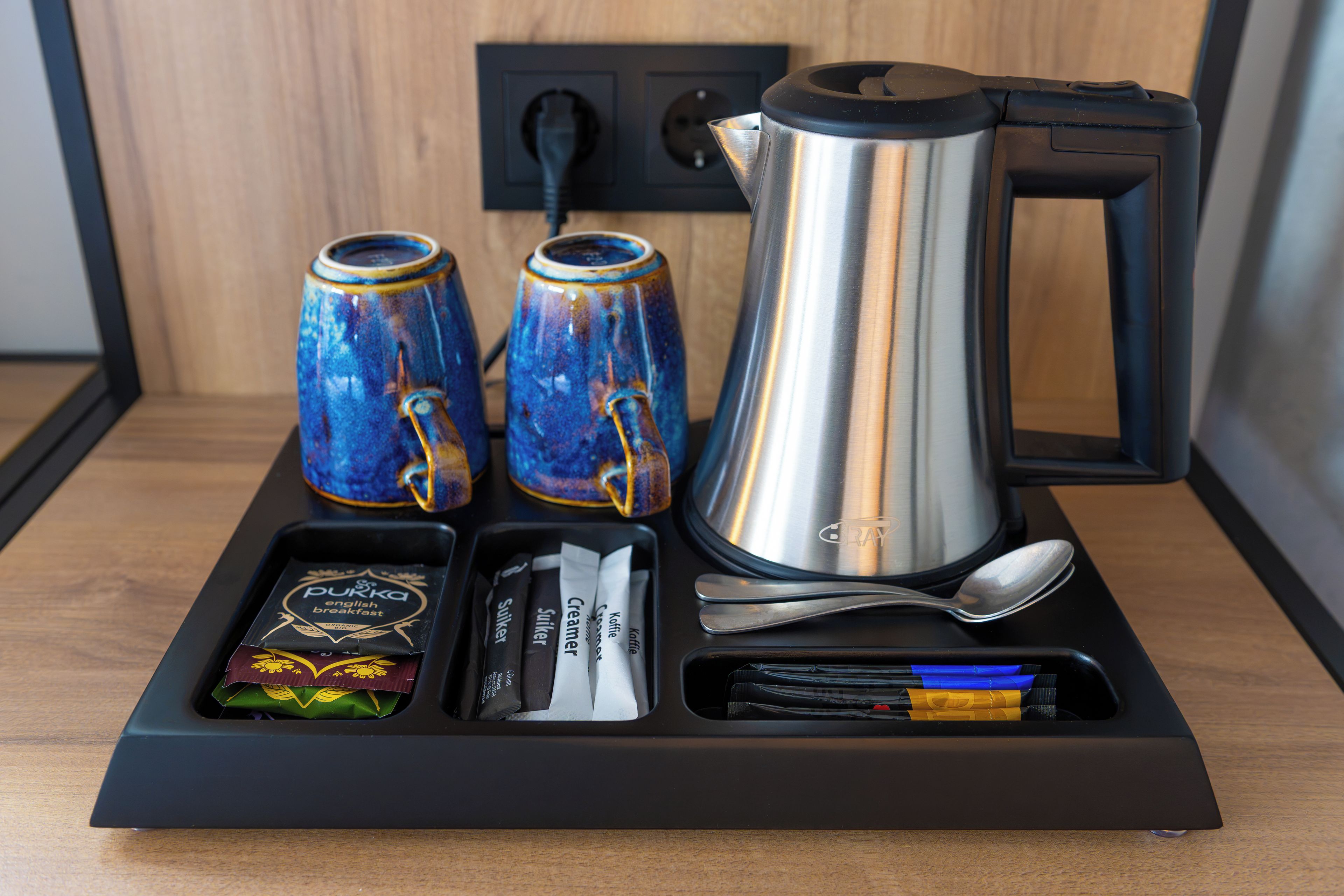 A coffee maker, two mugs, a spoon, a box of creamer, and a box of sugar packets on a tray.