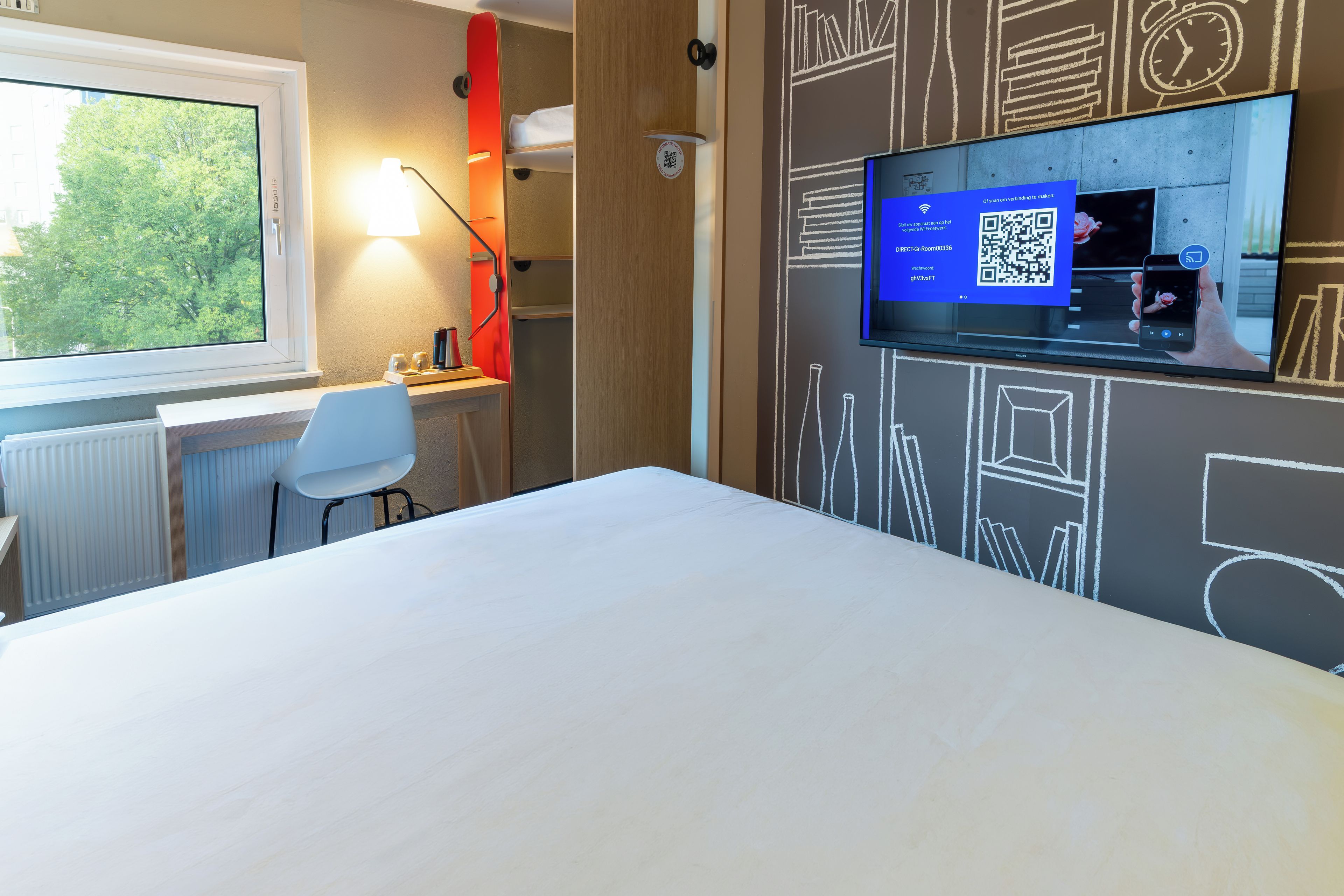 A hotel room with a bed, a desk with a chair, a window, and a wall-mounted TV displaying a QR code.